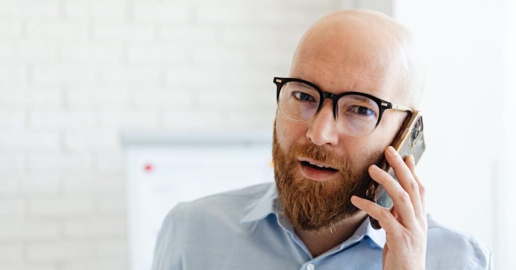Bald bearded man in glasses using smartphone inside an office setting.
